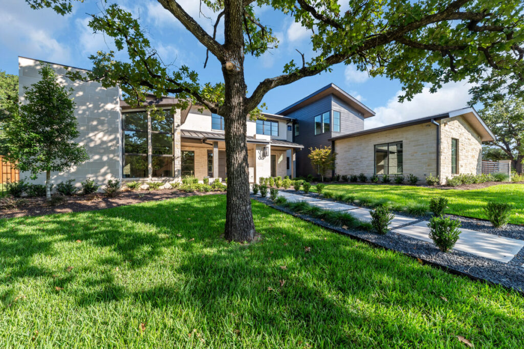 Modern custom home exterior with limestone and stucco detailing, manicured lawn, mature trees, and a curved stone walkway leading to the front entry on a landscaped residential lot.