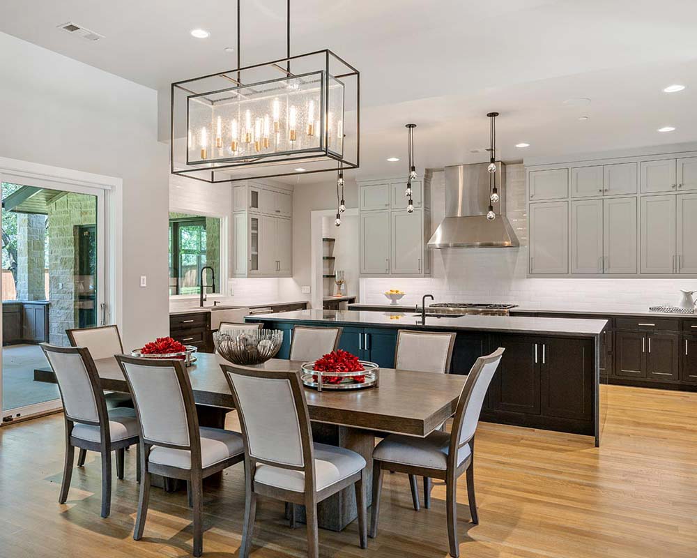 Open-concept custom kitchen featuring white cabinetry, a dark wood island, stainless steel range hood, and a dining table positioned beneath a rectangular linear chandelier.