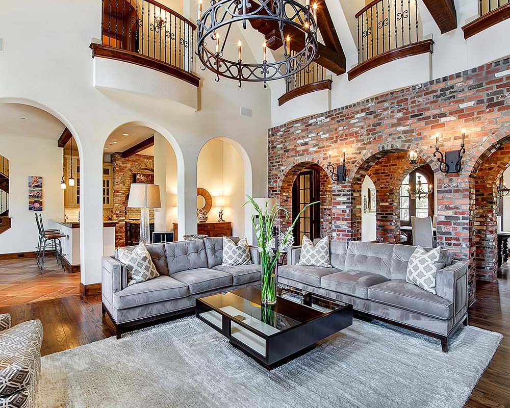 Two-story living room with exposed brick archways, wood flooring, a central seating arrangement, and a circular chandelier connecting the main level to the balcony above.