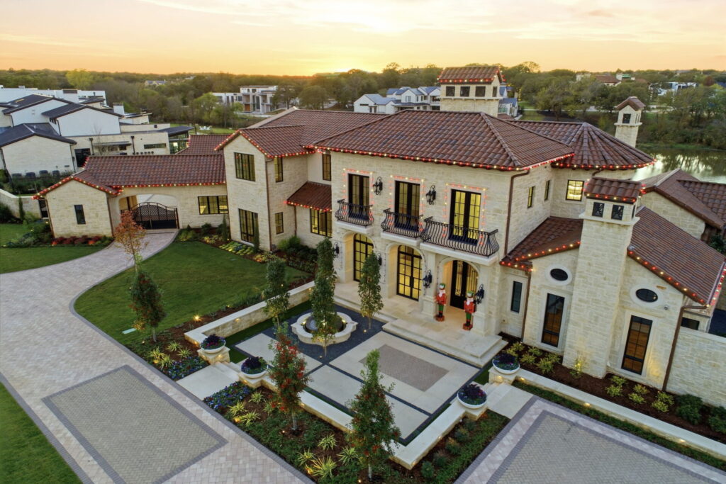 Custom Built Luxury Home Aerial dusk view of the estate's front entry and facade with warm exterior lighting, a circular motor court approach, and the lake visible beyond the property
