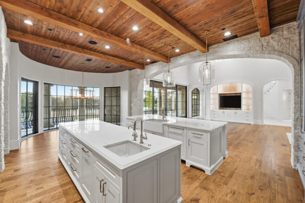 Open-concept custom kitchen with exposed wood ceiling beams, dual islands with light cabinetry, hardwood floors, and large windows bringing in natural light.