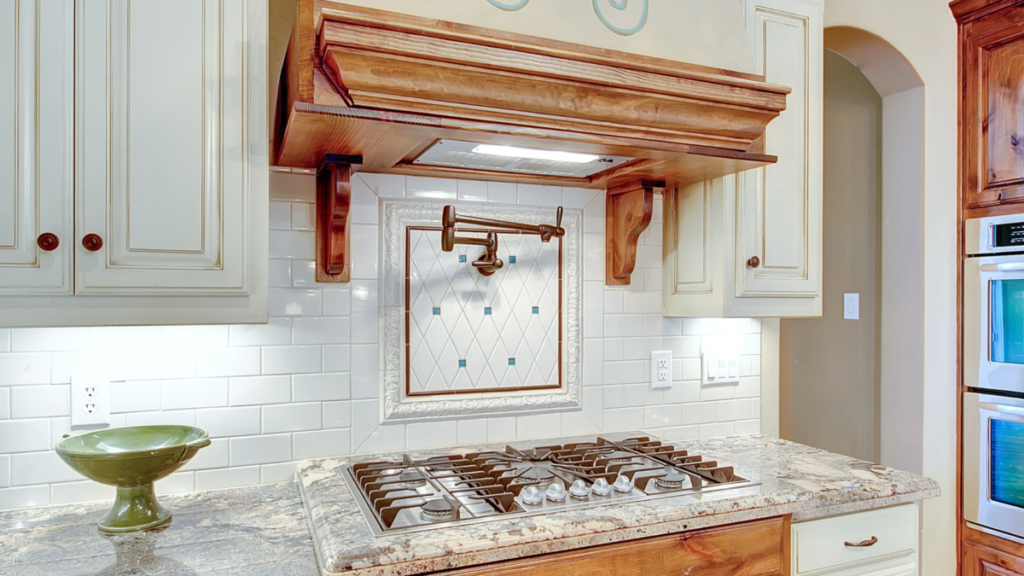 Traditional wall-mounted pot filler installed above a gas cooktop in a custom kitchen with wood cabinetry, decorative tile backsplash, and stone countertops.