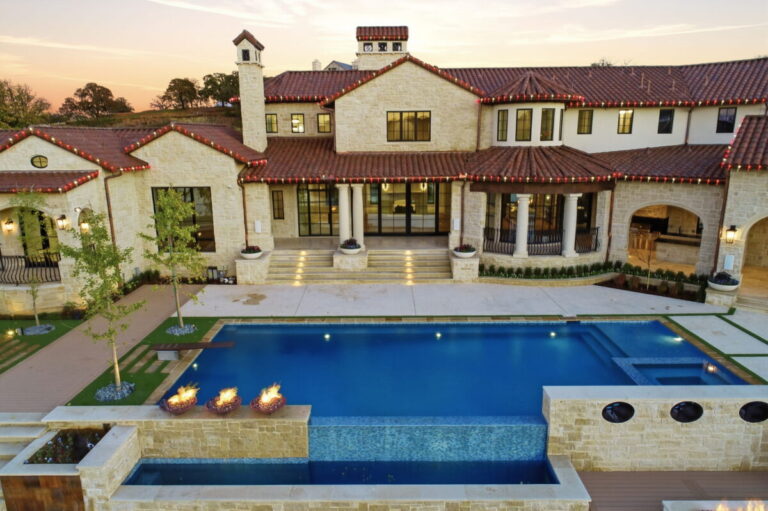 Aerial daytime view of the rear of the Mediterranean estate showing the pool, covered outdoor loggia, and landscaped backyard grounds from above