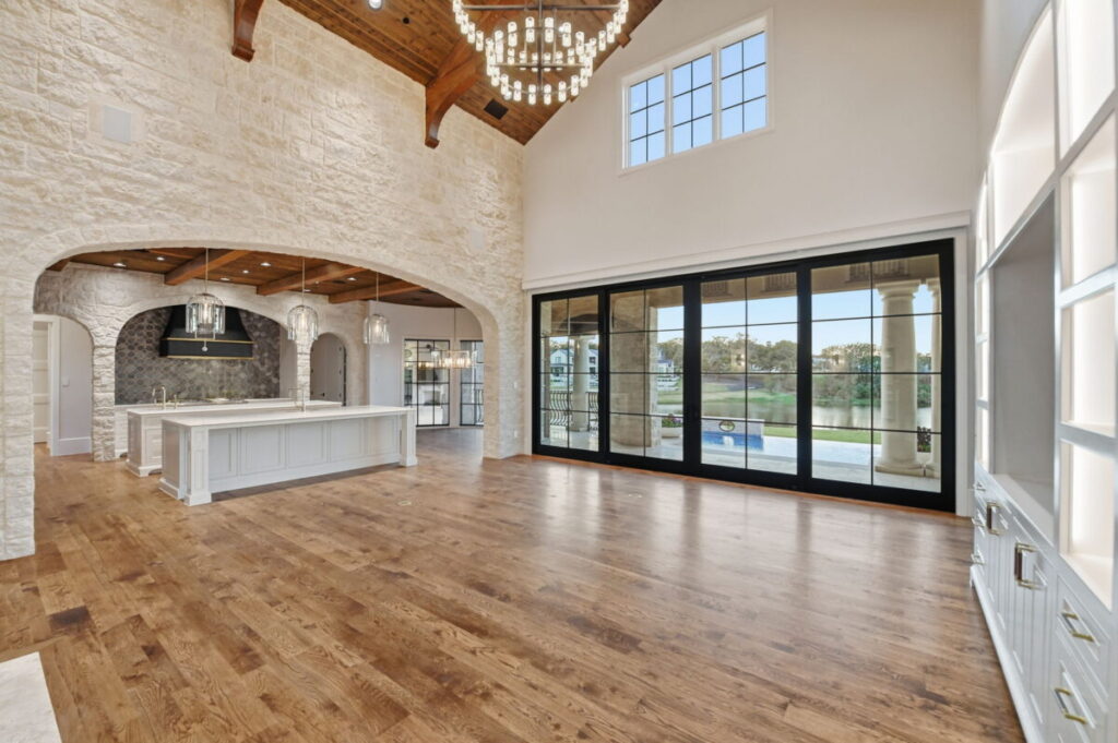 Open-concept living room in a custom luxury home featuring wide-plank hardwood floors, vaulted ceilings with exposed wood beams, a stone fireplace, large sliding glass doors, and a view into a gourmet kitchen with a large island.