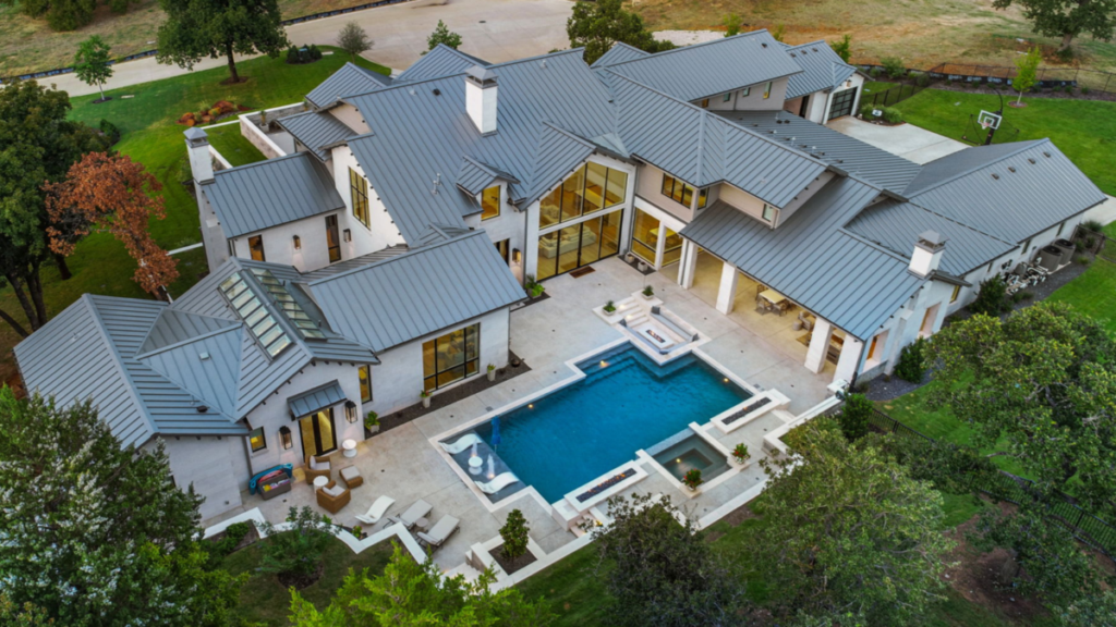 Aerial view of a large custom luxury home with a standing seam metal roof, expansive backyard patio, swimming pool with spa, covered outdoor living areas, and landscaped grounds surrounded by mature trees.