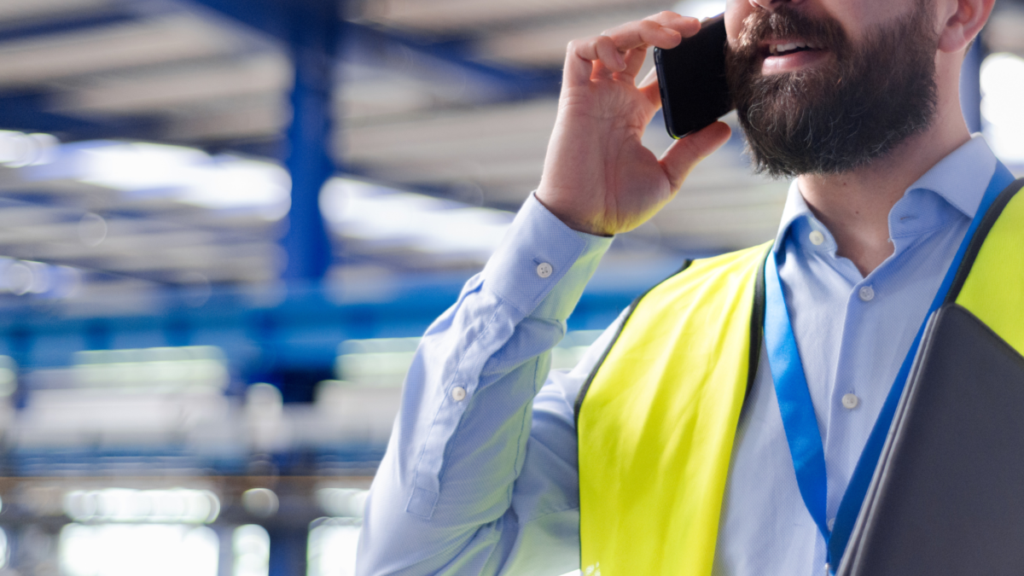 Construction professional wearing a high-visibility safety vest speaking on a mobile phone while holding a clipboard inside a warehouse or industrial facility.
