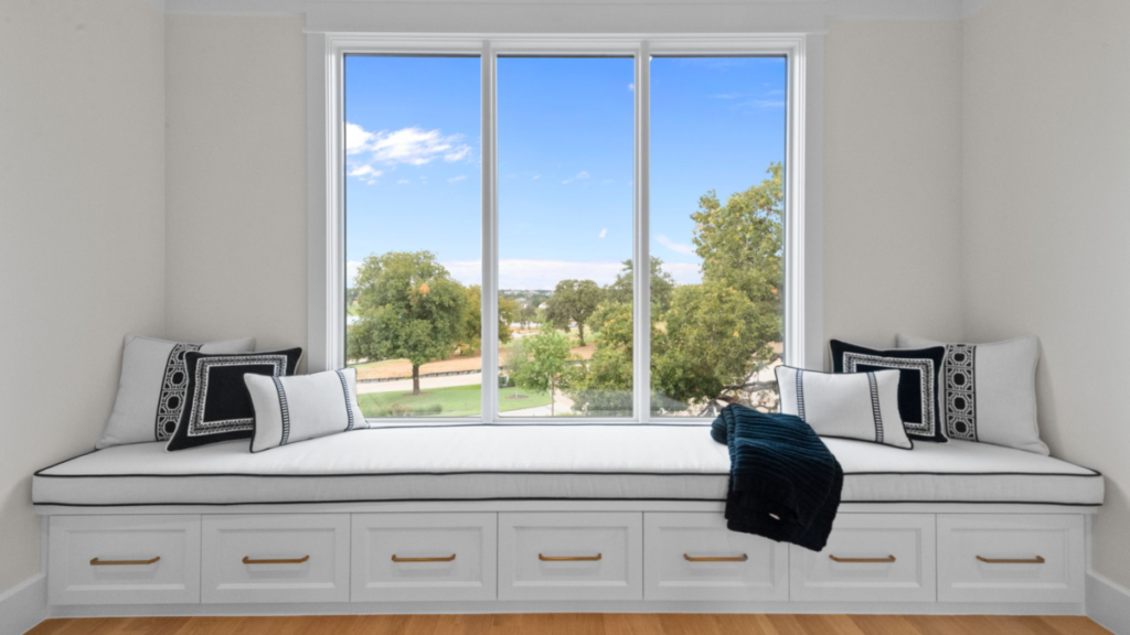 Built-in window bench with white cabinetry drawers and black-and-white accent pillows overlooking a landscaped outdoor view through large windows.