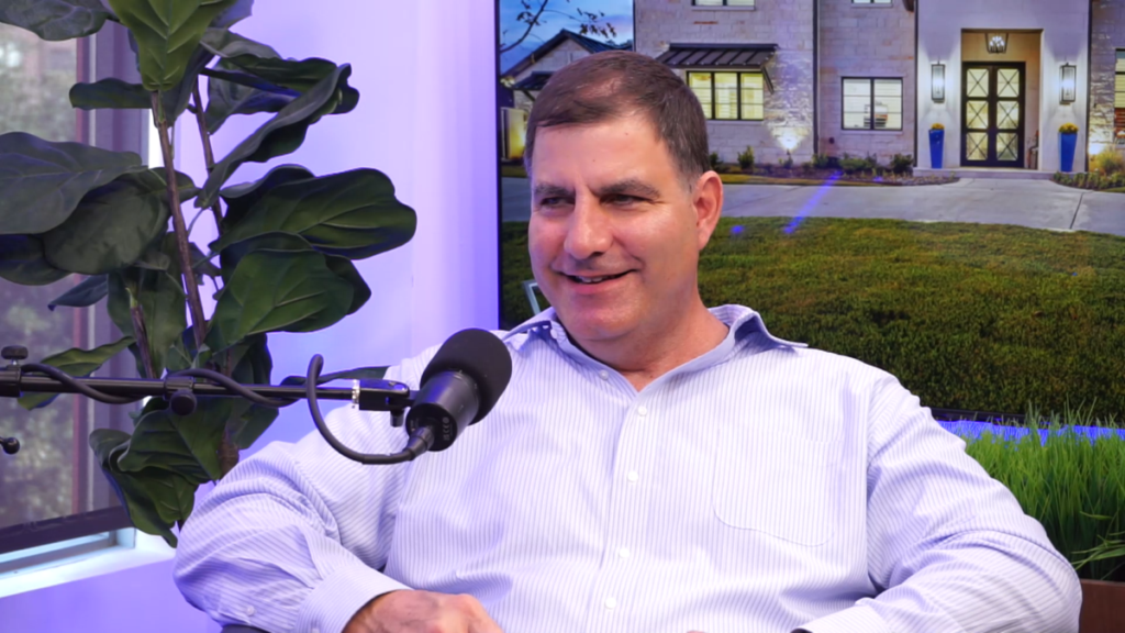 Mike Garabedian of Garabedian Properties smiling on the podcast set of Uniqueness... "Built-In" in Southlake, Texas wearing a blue button-up with a microphone in front of him.