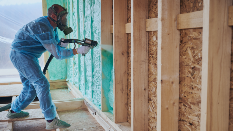 Installer wearing a respirator and protective suit spraying green foam insulation inside a framed residential wall system.