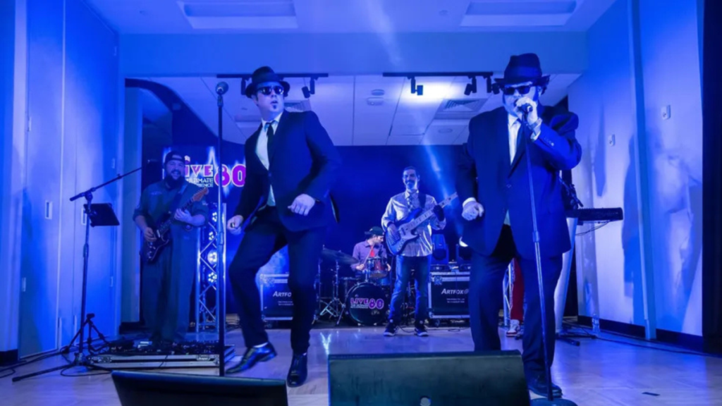 Two Blues Brothers tribute performers in black suits, fedoras, and sunglasses command the front of a stage, dancing and singing into microphones while a live band plays behind them, the entire scene awash in electric blue stage lighting.