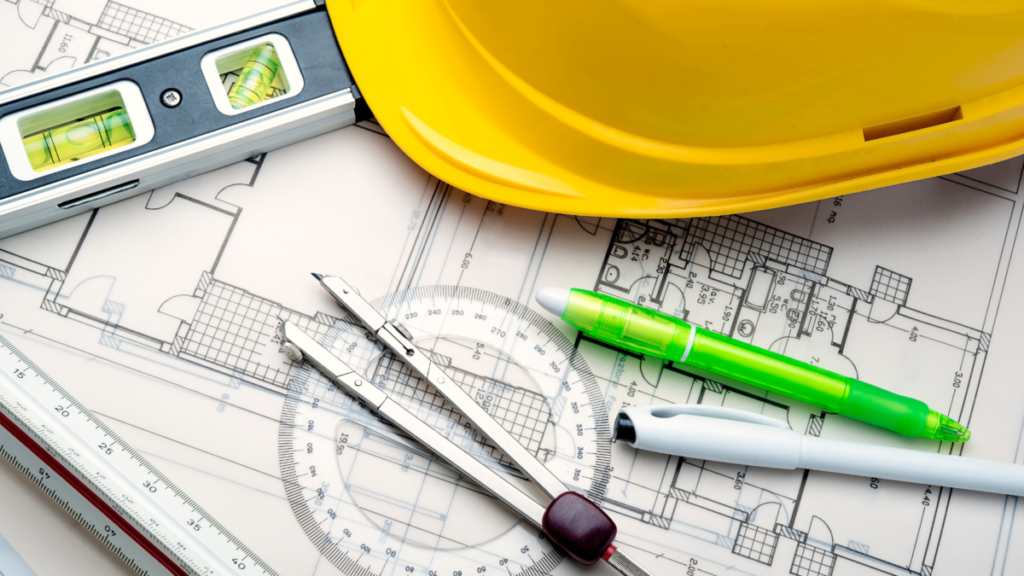 Architectural blueprints spread across a drafting table alongside a compass, protractor, ruler, green highlighter, and yellow hard hat, representing the precision planning behind custom home construction.