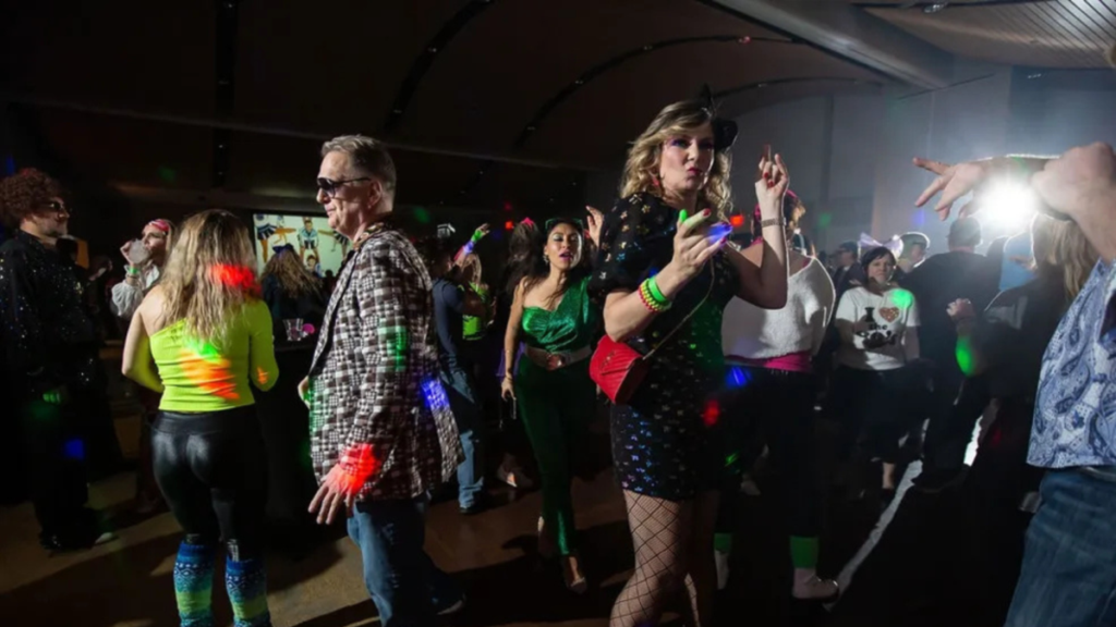 A packed dance floor at The Retro Dance Party, with costumed guests dancing beneath multicolored neon lights — neon green leg warmers, sequined outfits, and glowing wristbands visible throughout the exuberant crowd, with music videos projected on a screen in the background.