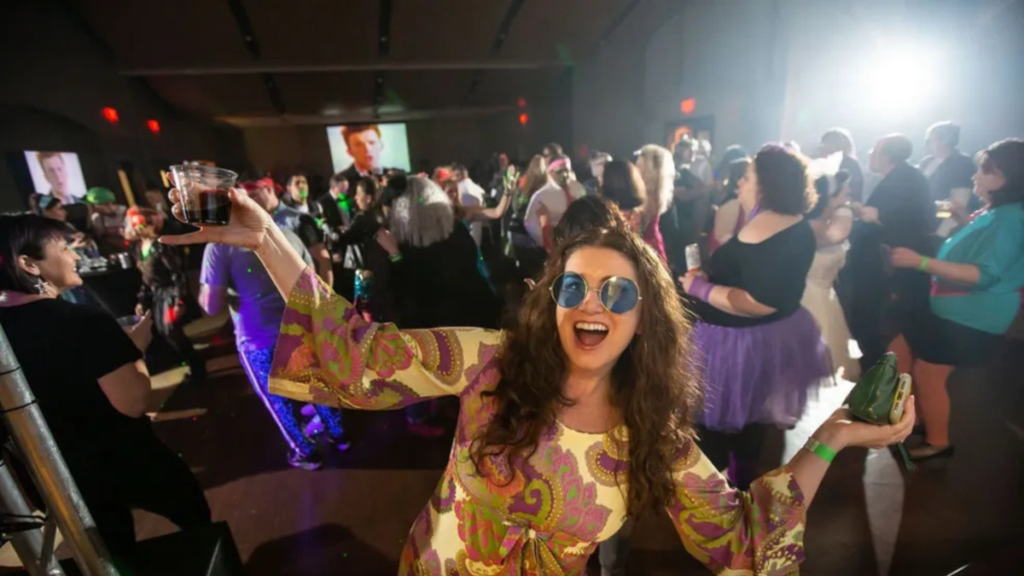 A woman in a colorful paisley dress and round blue sunglasses raises her drink to the crowd at The Retro Dance Party, beaming with elation as costumed dancers fill the floor behind her and music videos glow on large screens throughout the venue.