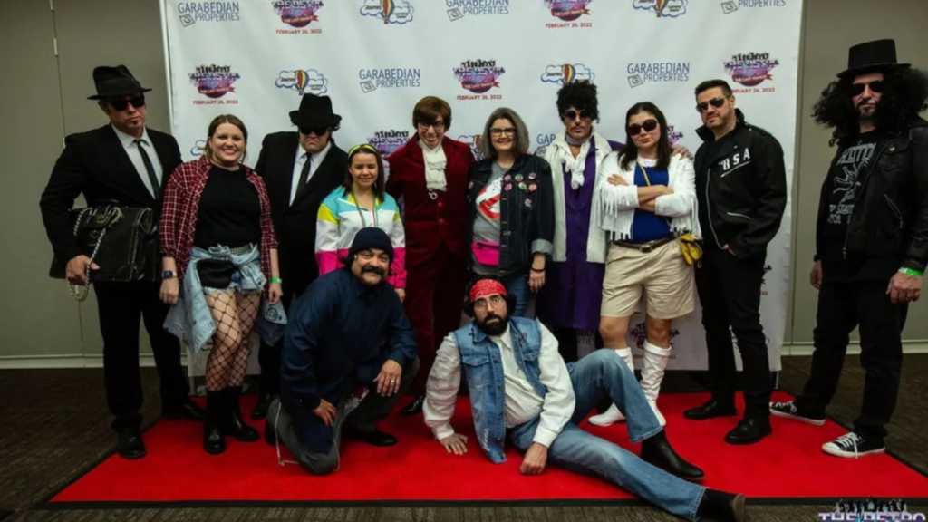 A large group of costumed guests poses on the red carpet at The Retro Dance Party in front of a Garabedian Properties step-and-repeat backdrop, dressed as iconic characters including the Blues Brothers, Austin Powers, Cheech and Chong, and various retro pop culture figures.