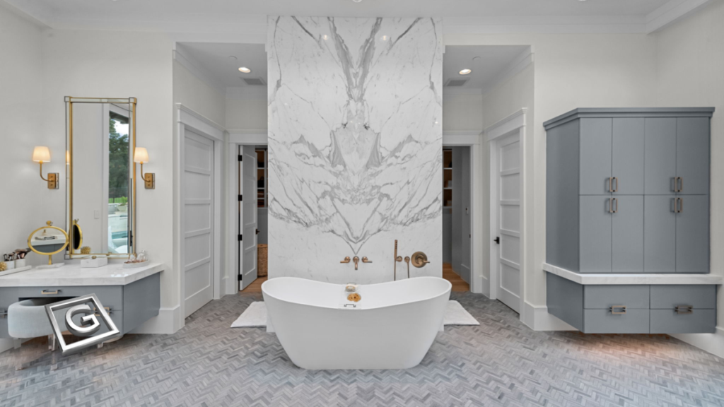 Garabedian Properties artisan home construction specialist North Texas — spa-inspired primary bathroom with freestanding soaking tub, floor-to-ceiling vein-matched marble accent wall, herringbone mosaic tile flooring, floating gray vanity cabinetry, and brushed gold wall-mounted fixtures