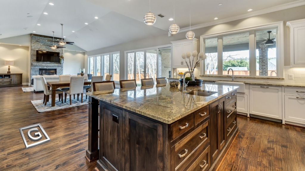 Garabedian Properties luxury custom home builder Southlake Texas — open-concept kitchen and living space featuring a large dark walnut island with granite countertop, layered pendant and recessed lighting, stacked stone fireplace, and expansive windows with views to the backyard