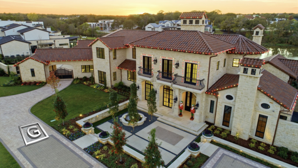 Garabedian Properties premier residential builder Westlake Texas — aerial twilight view of a completed Mediterranean-style custom luxury home featuring a clay tile roof, natural limestone exterior, wrought iron balconies, arched entry, manicured courtyard with circular fountain, and holiday lighting throughout, situated adjacent to a private pond