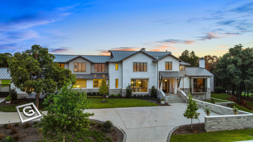 Garabedian Properties luxury custom home builder Keller Texas — dusk aerial view of a completed modern farmhouse custom home featuring a white stucco and board-formed concrete exterior, standing seam metal roof, expansive black-framed windows, stone retaining walls, mature oak trees, and a circular drive illuminated by landscape lighting