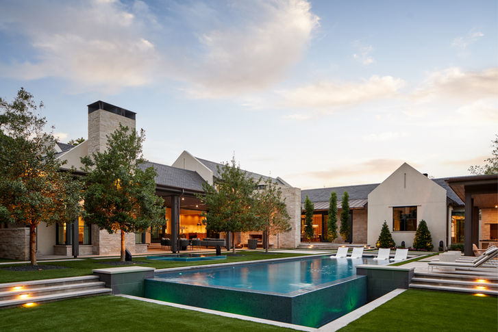 Resort-style pool and landscaped outdoor living space at dusk, designed by Osterberger Design Group. Artisan home construction with stone and white exterior, mature trees, and illuminated interiors.