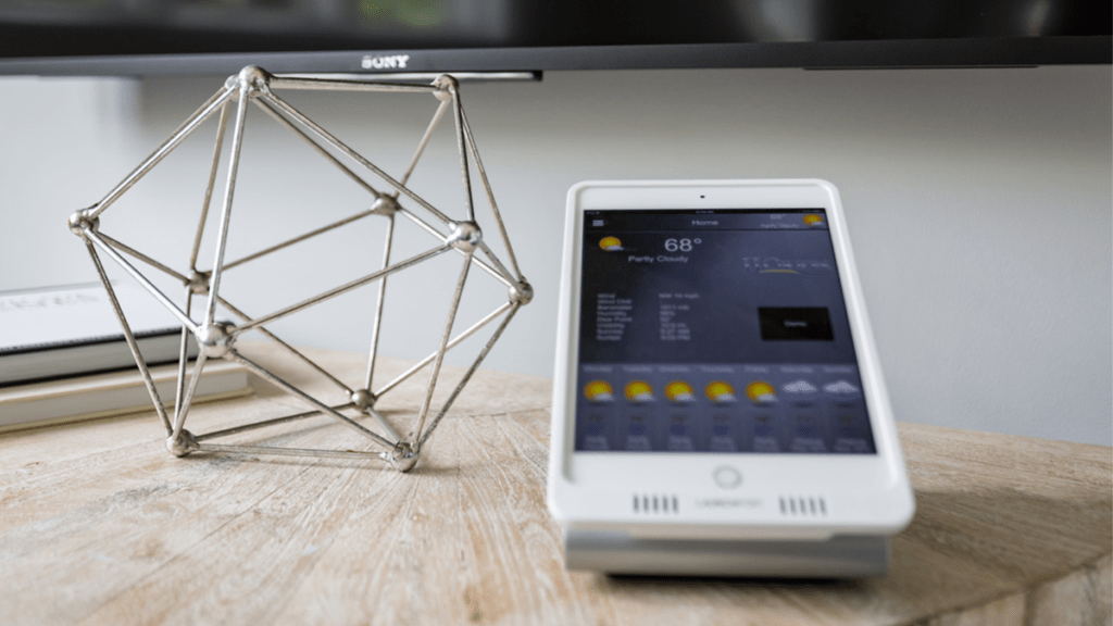 Smart home control tablet displaying weather forecast and current temperature, mounted on a stand beside a geometric metal sculpture on a wooden desk with a television in the background.