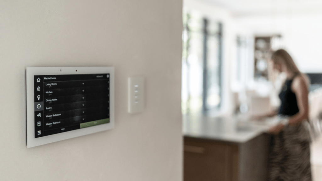 Wall-mounted touchscreen control panel displaying a media zones list for living room, kitchen, dining room, pantry, master bathroom, and master bedroom, with a blurred figure visible in a kitchen beyond.