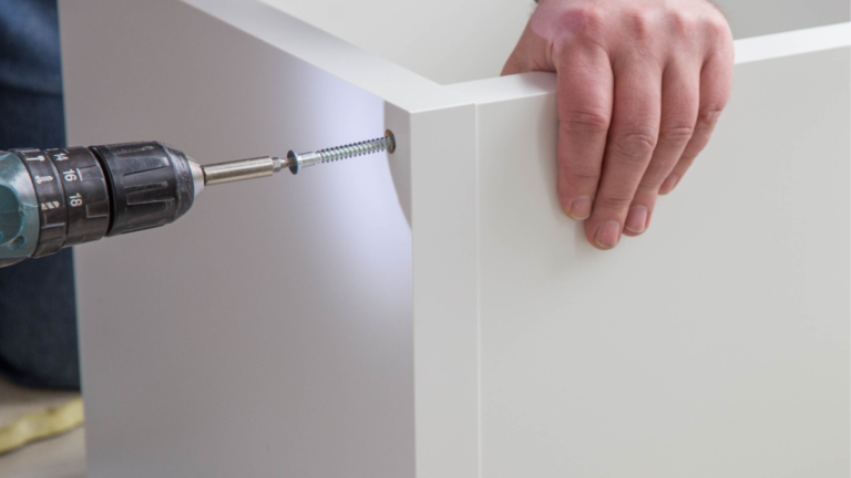 Close-up of a power drill driving a screw into the corner joint of a white flat-pack furniture panel, with one hand steadying the piece from the opposite side.