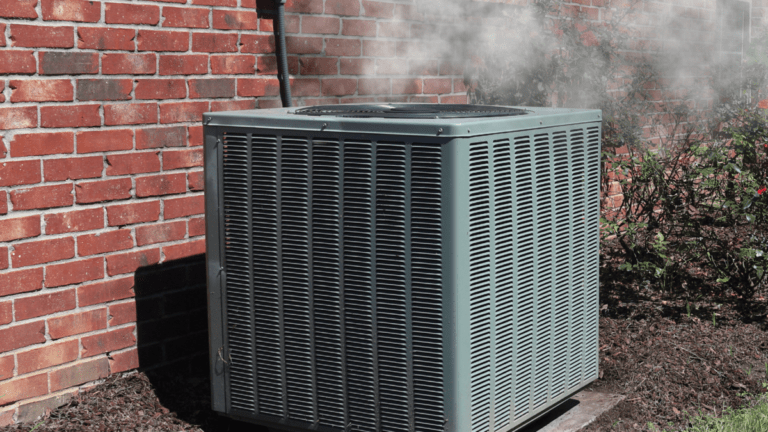 An outdoor air conditioning condenser unit against a red brick exterior wall, with a cloud of white vapor rising from its top fan grille. Rose bushes and greenery surround the unit, with dappled sunlight visible on the brick.