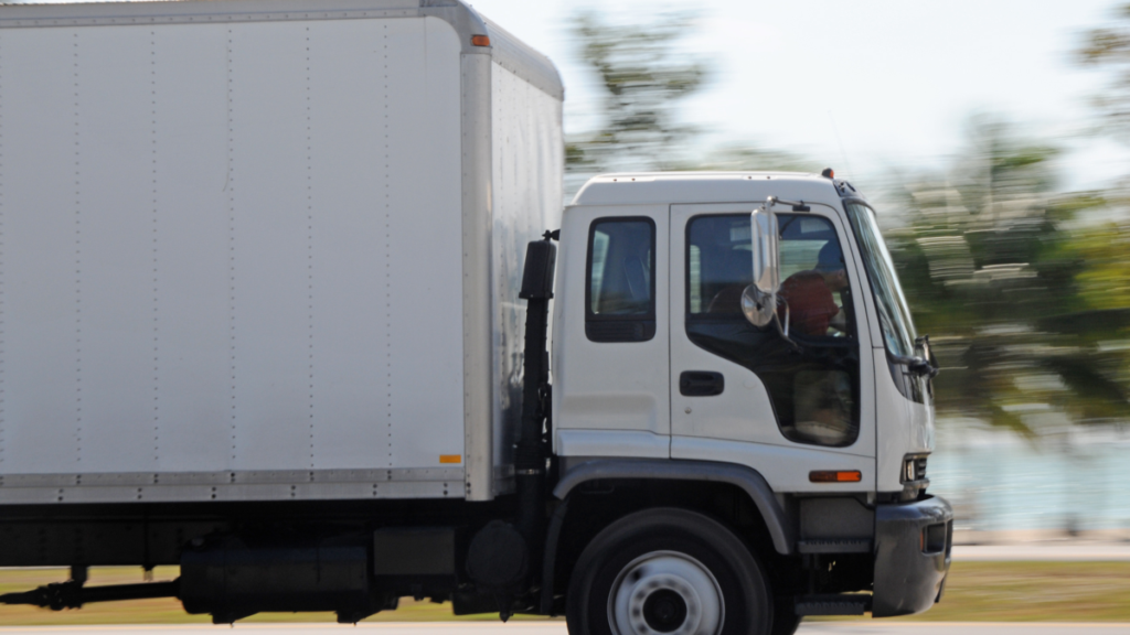A white box truck in motion on an open road, with trees blurred in the background.