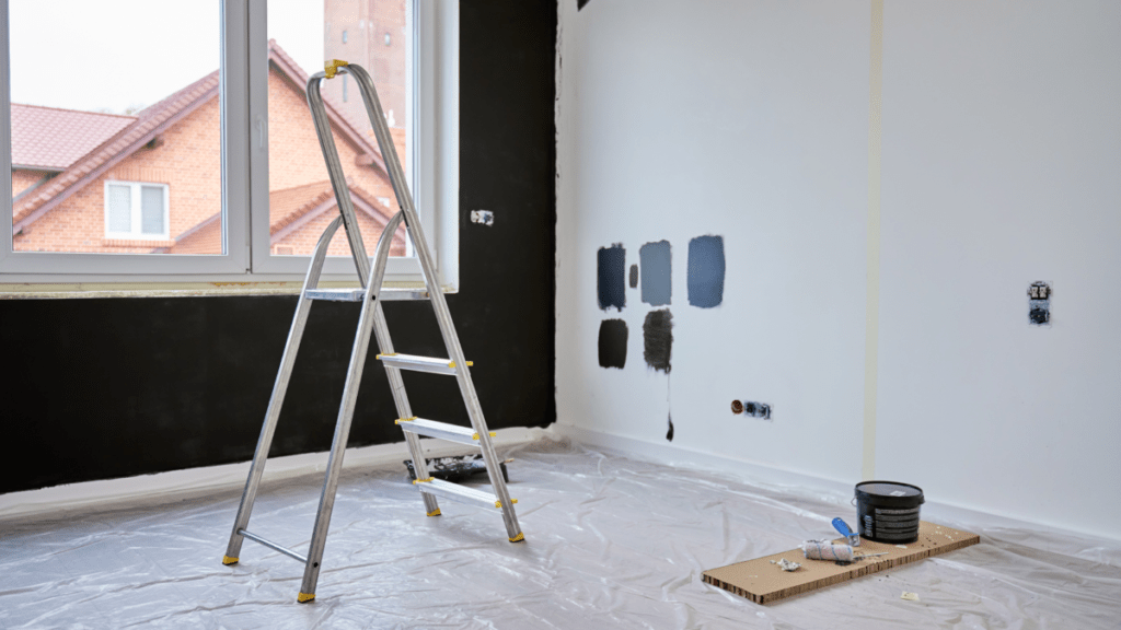 An empty room during a custom home build with a step ladder, a plastic drop cloth covering the floor, and several dark blue and gray paint swatches brushed onto the wall for comparison.
