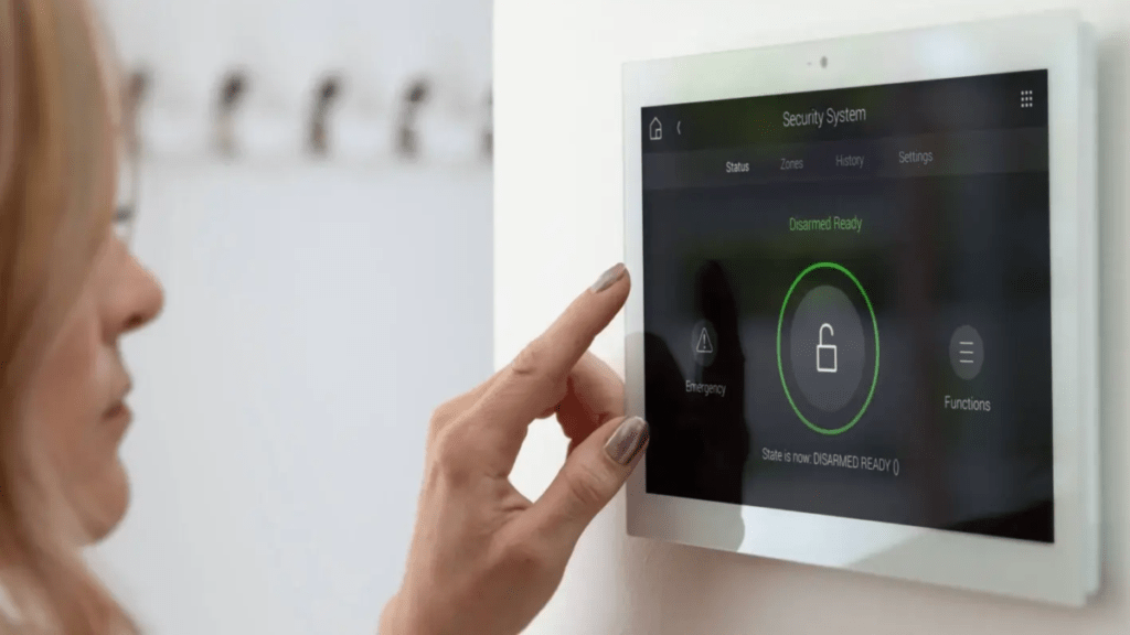 Woman touching a wall-mounted security system touchscreen displaying "Disarmed Ready" status with a green unlock icon, emergency and functions buttons on either side.