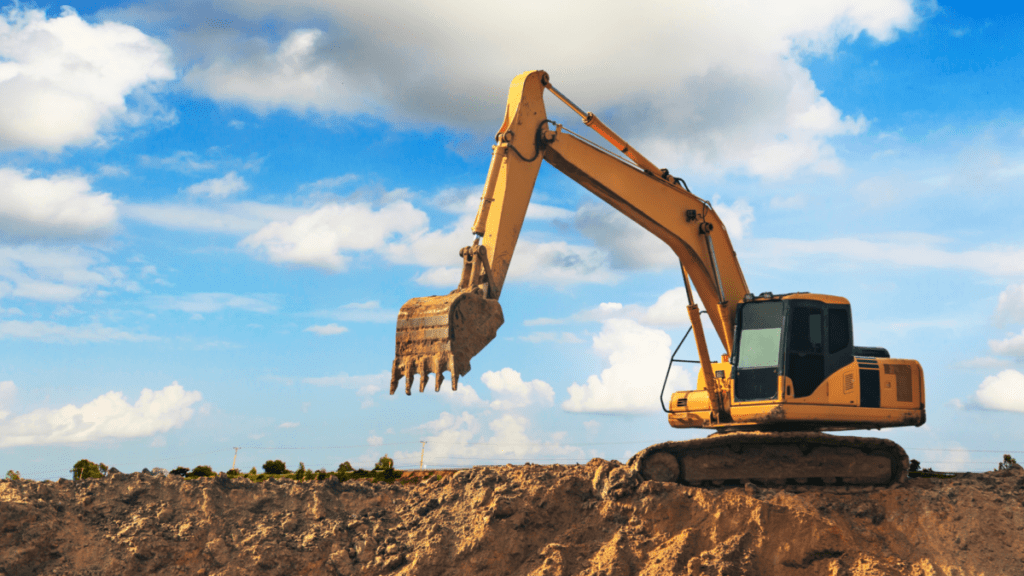 Skid steer loader with a raised bucket dumping loose soil into an open excavation beside a residential lawn, dust drifting from the falling material. String line and a marker flag define the edge of the dig, with manicured grass, shrubs, and decorative rock beds visible in the background.