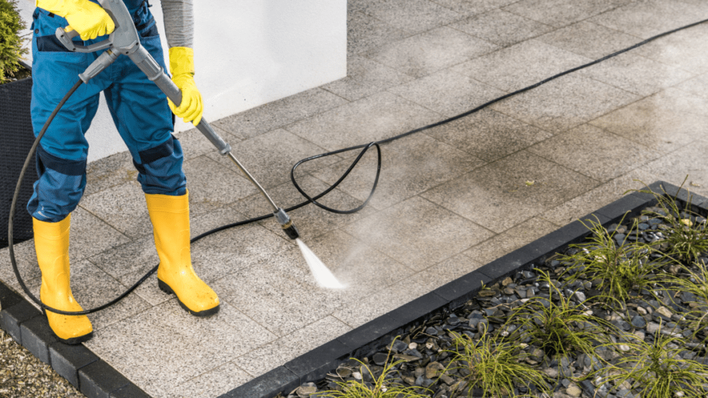 Worker in yellow gloves and boots power washing a stone paver patio along the base of a home exterior, with a planting bed protected nearby.