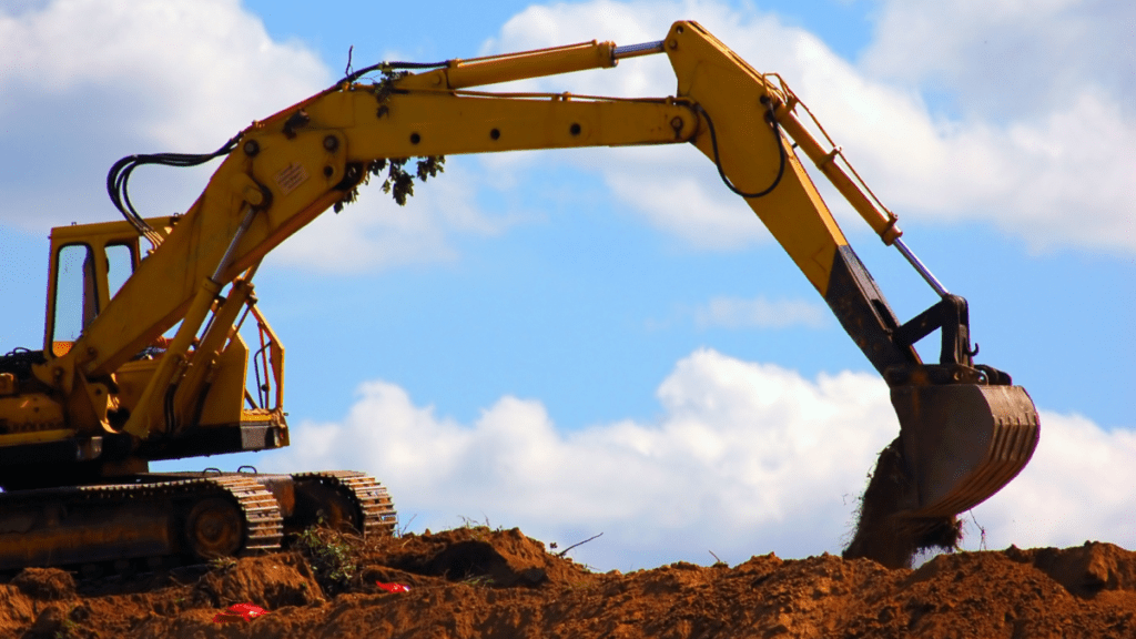 Yellow tracked excavator viewed against a bright blue sky with scattered clouds, arm fully extended to the right as the bucket lifts a load of soil with roots and vegetation trailing from its edge. Reddish brown earth and scattered green growth cover the working surface below the machine.