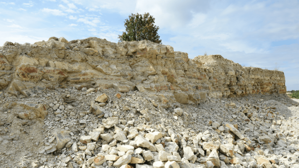 Exposed limestone formation cut into a hillside, showing horizontal bedding planes in cream and rust colored bands. Broken pale stone rubble in various sizes covers the slope in the foreground, and a single leafy tree stands at the top of the ridge against a partly cloudy sky.