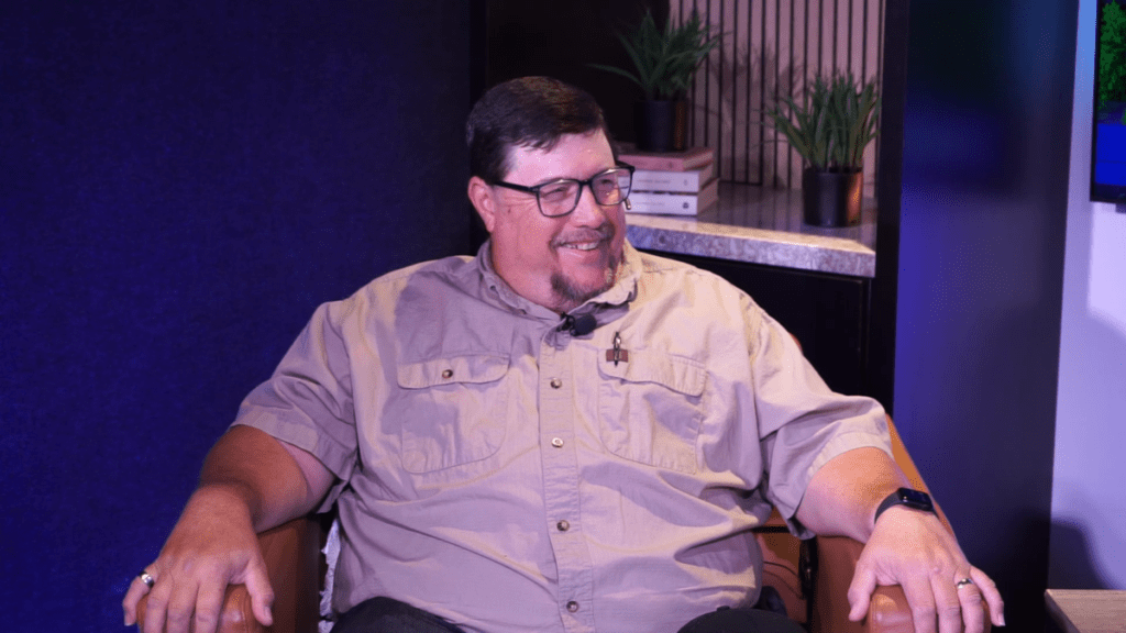 Mike, owner operator of Mike's Trees and More, seated in a leather chair and smiling off camera during a recorded episode of Uniqueness... "Built-In". He wears a tan button-down work shirt with a clip-on microphone, and the set behind him features a deep blue acoustic wall, potted plants, and stacked books on a stone counter.