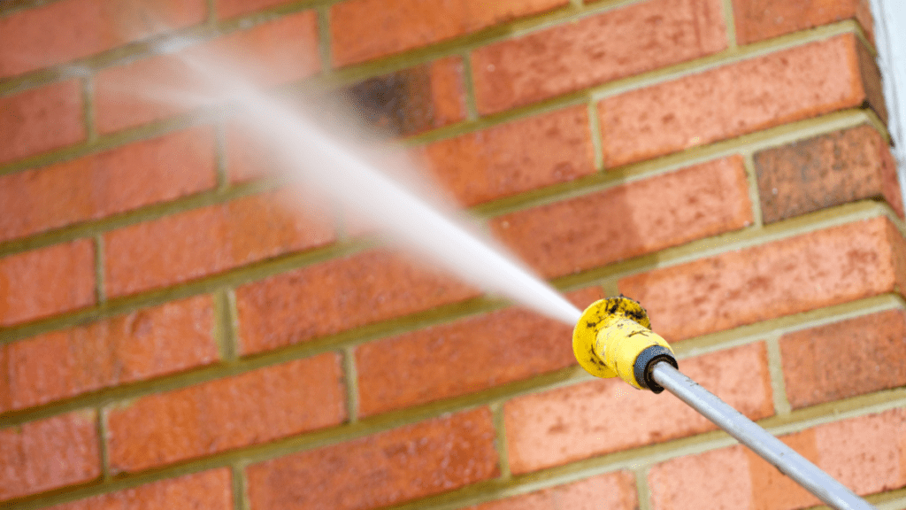 Pressure washer nozzle spraying a fan of water across a red brick wall, cleaning the brick face and mortar joints of accumulated soil.