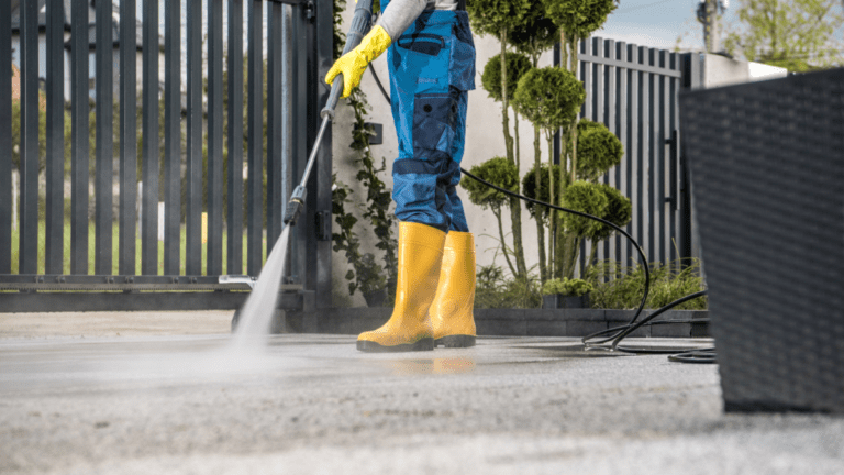 Worker power washing a concrete driveway beside an iron entry gate and manicured hedges as part of annual exterior home maintenance.