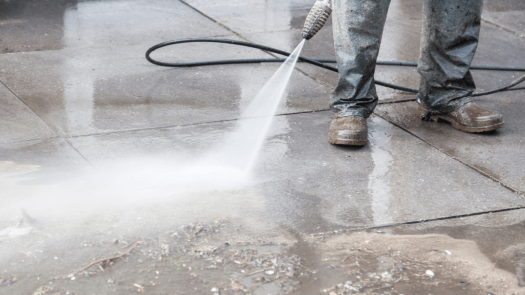 Pressure washer wand clearing dirt and organic debris from a concrete patio, demonstrating the fan spray pattern used for hard surfaces.