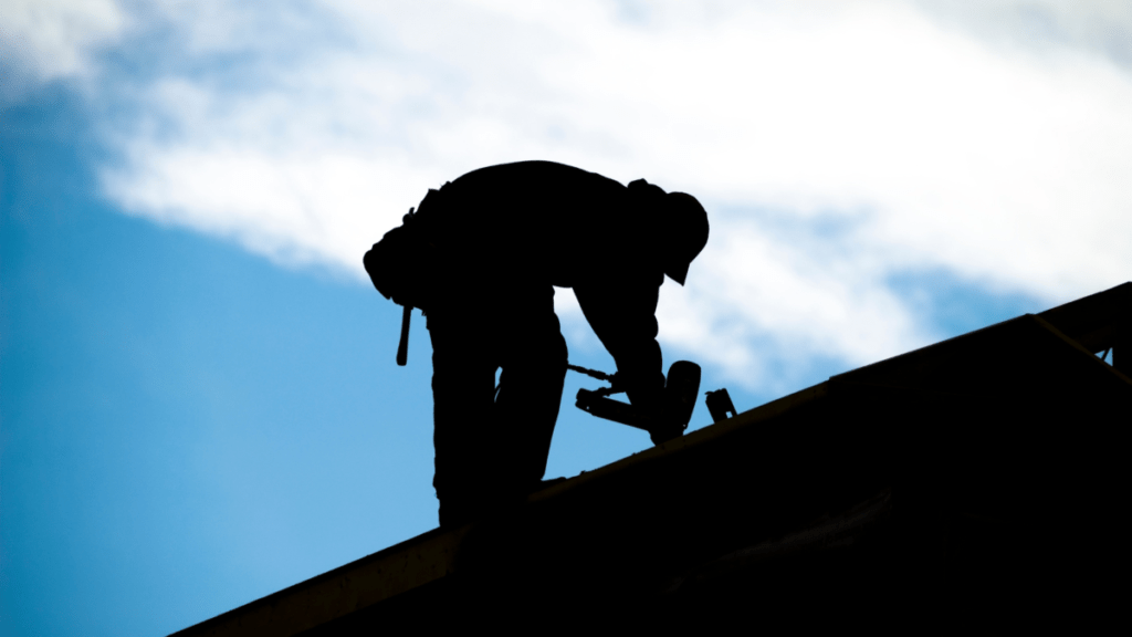 Silhouette of a roofing professional kneeling on a rooftop against a bright blue sky, using a nail gun to secure shingles.