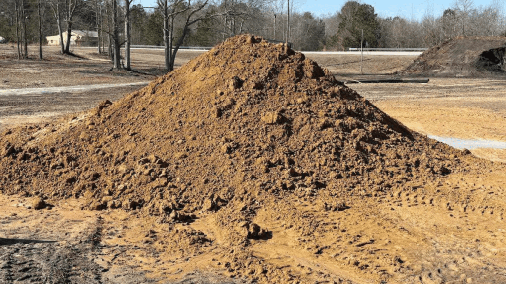 Cone-shaped stockpile of tan sandy clay fill sitting on a cleared and graded site, with tire tracks pressed into the surrounding ground. Bare winter trees, a guardrail, and a distant building line the background under a clear sky.