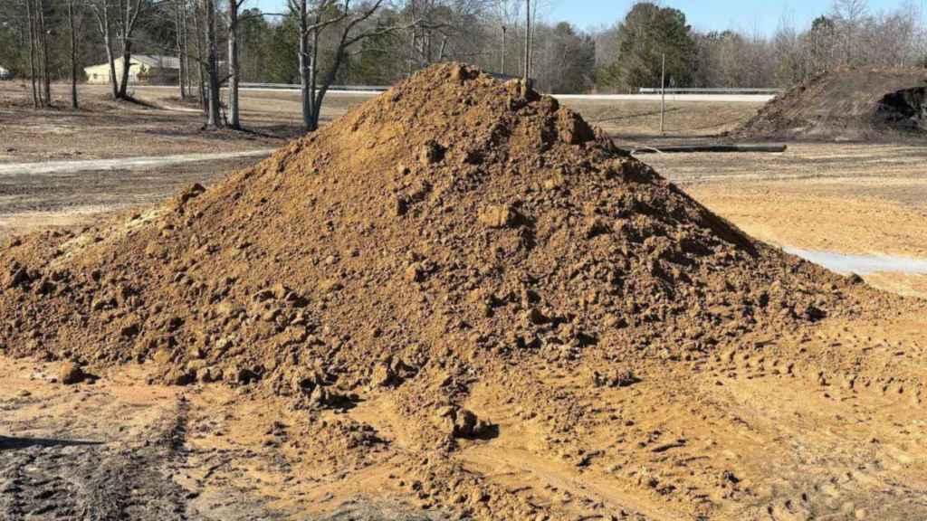 Cone-shaped stockpile of tan sandy clay fill sitting on a cleared and graded site, with tire tracks pressed into the surrounding ground. Bare winter trees, a guardrail, and a distant building line the background under a clear sky.