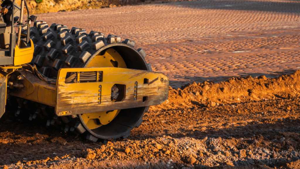 Close-up of a yellow sheepsfoot compactor with a knobbed steel drum working across a pad of reddish soil at golden hour. The compacted area behind the machine shows the regular pattern left by the drum feet, with loose material still to be worked in the foreground.