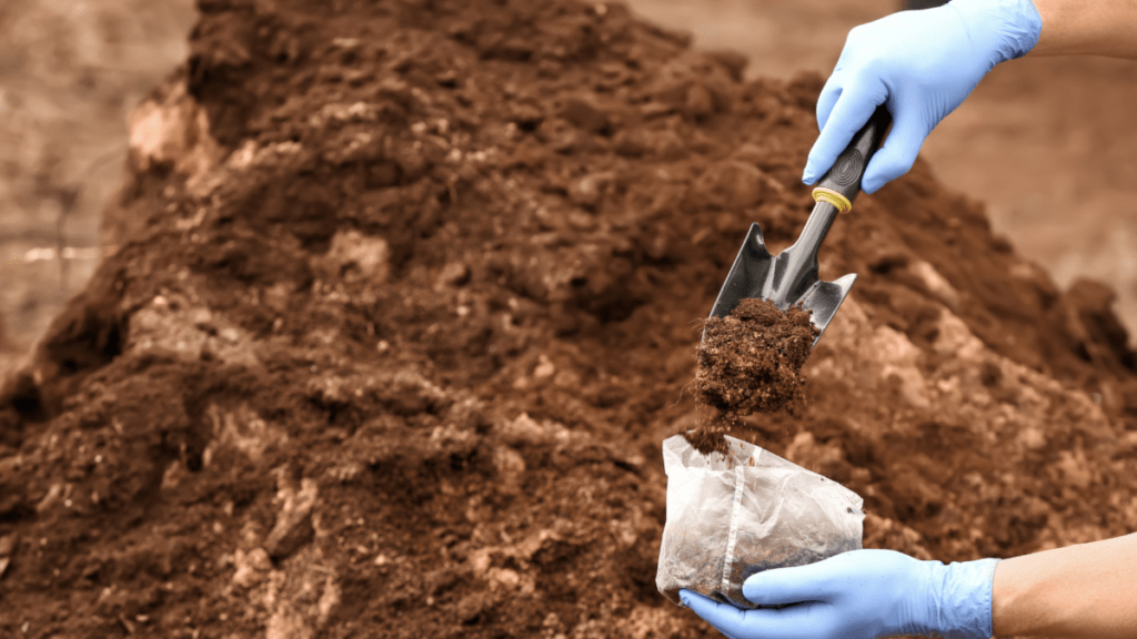 Gloved hands collecting a soil sample, using a small hand trowel to pour loose dark earth into a clear plastic collection bag. A large mound of brown soil fills the background, softly out of focus.