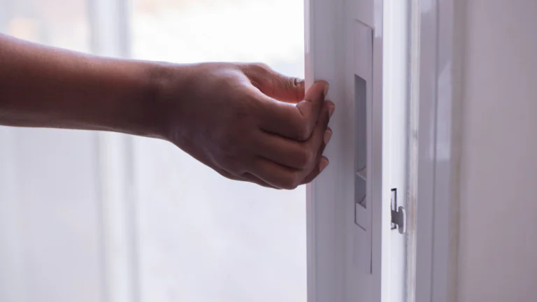 Close-up of a hand sliding open a white patio door, with a soft-focus bright window in the background and a metal handle and latch mechanism visible on the frame.