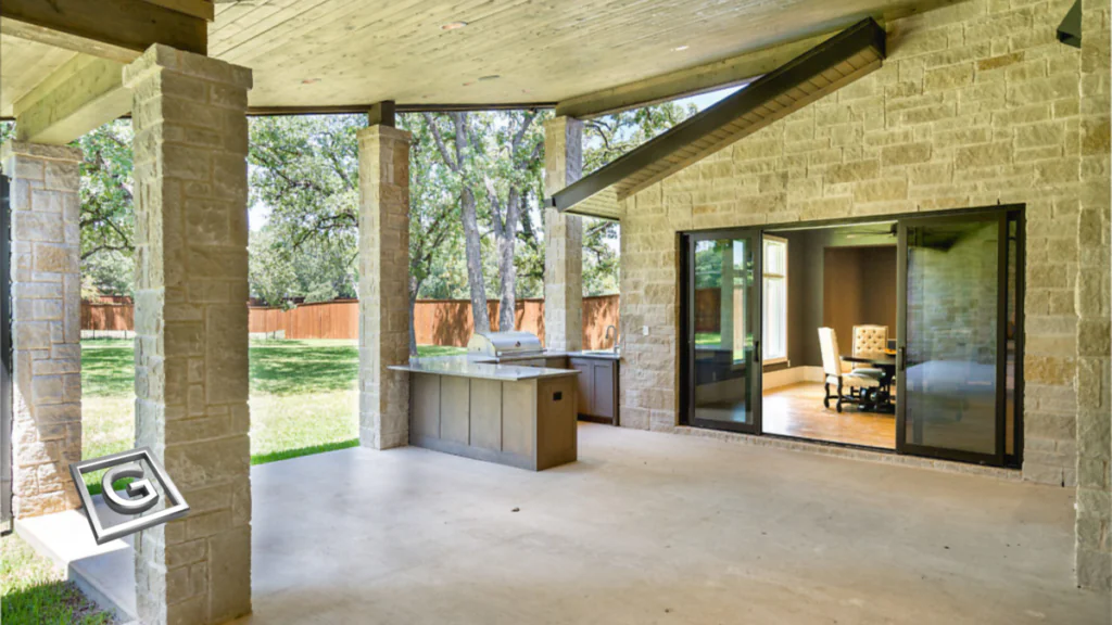 Covered stone patio with an outdoor kitchen featuring a built-in grill and counter, stone columns, and glass sliding doors opening into an interior dining room, with a backyard lawn and wood fence beyond.