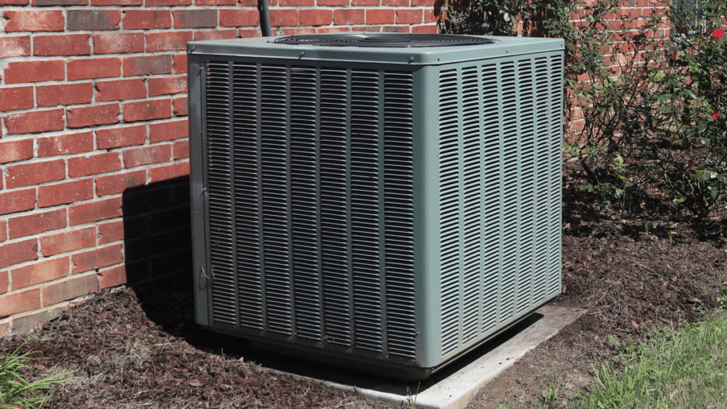 Weathered gray outdoor air conioning condenser unit positioned against a red brick wall, surrounded by mulch and a rose bush.